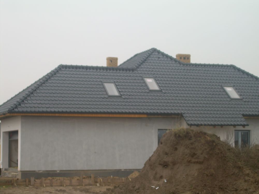 Dom jednorodzinny z grafitową dachówką, trzema oknami dachowymi, surową elewacją i stertą ziemi na pierwszym planie.